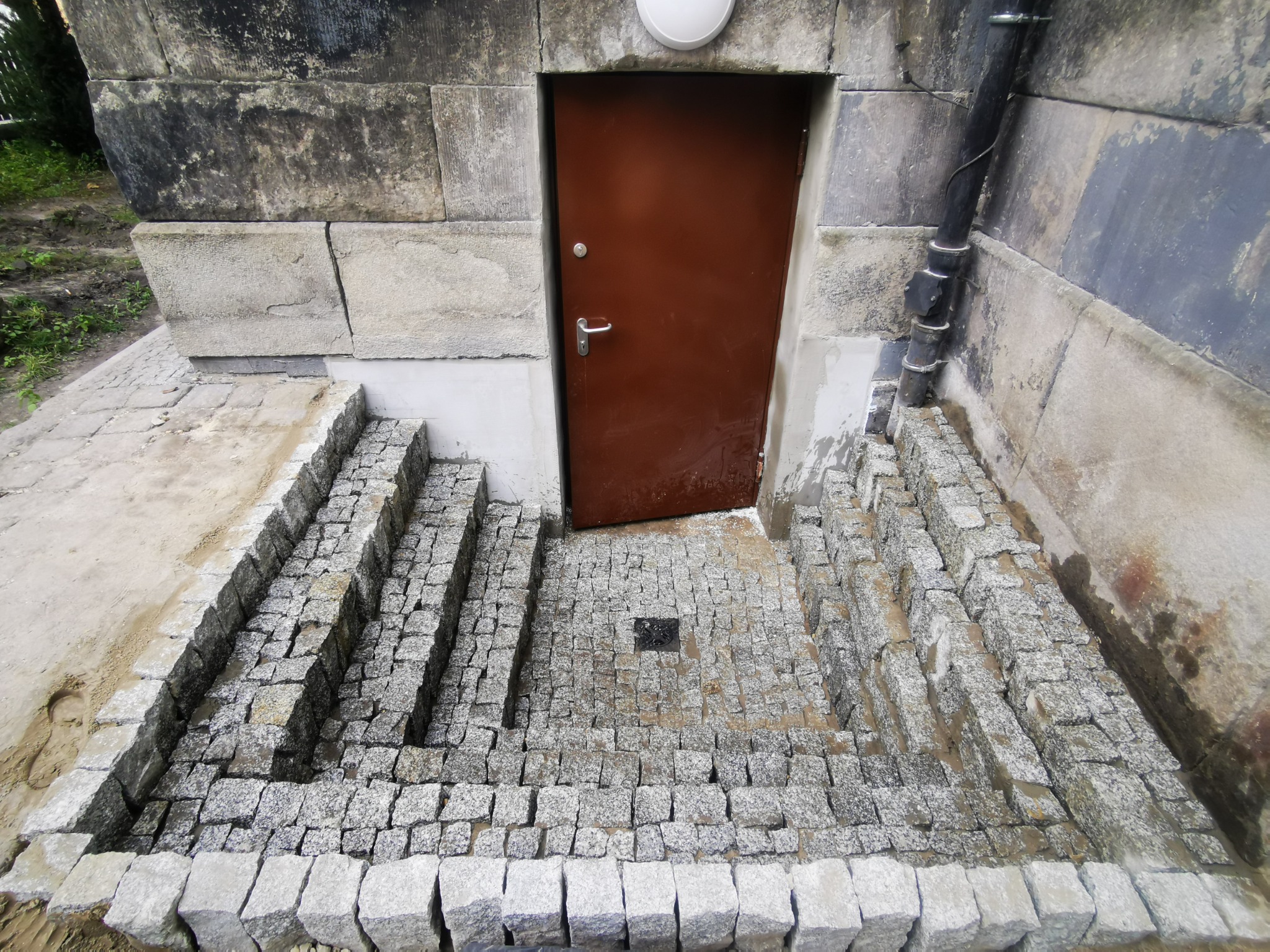 Schody wejściowe do budynku wyłożone szarą kostką granitową, prowadzące do brązowych drzwi wejściowych z metalową klamką, widoczne elementy elewacji z kamienia.