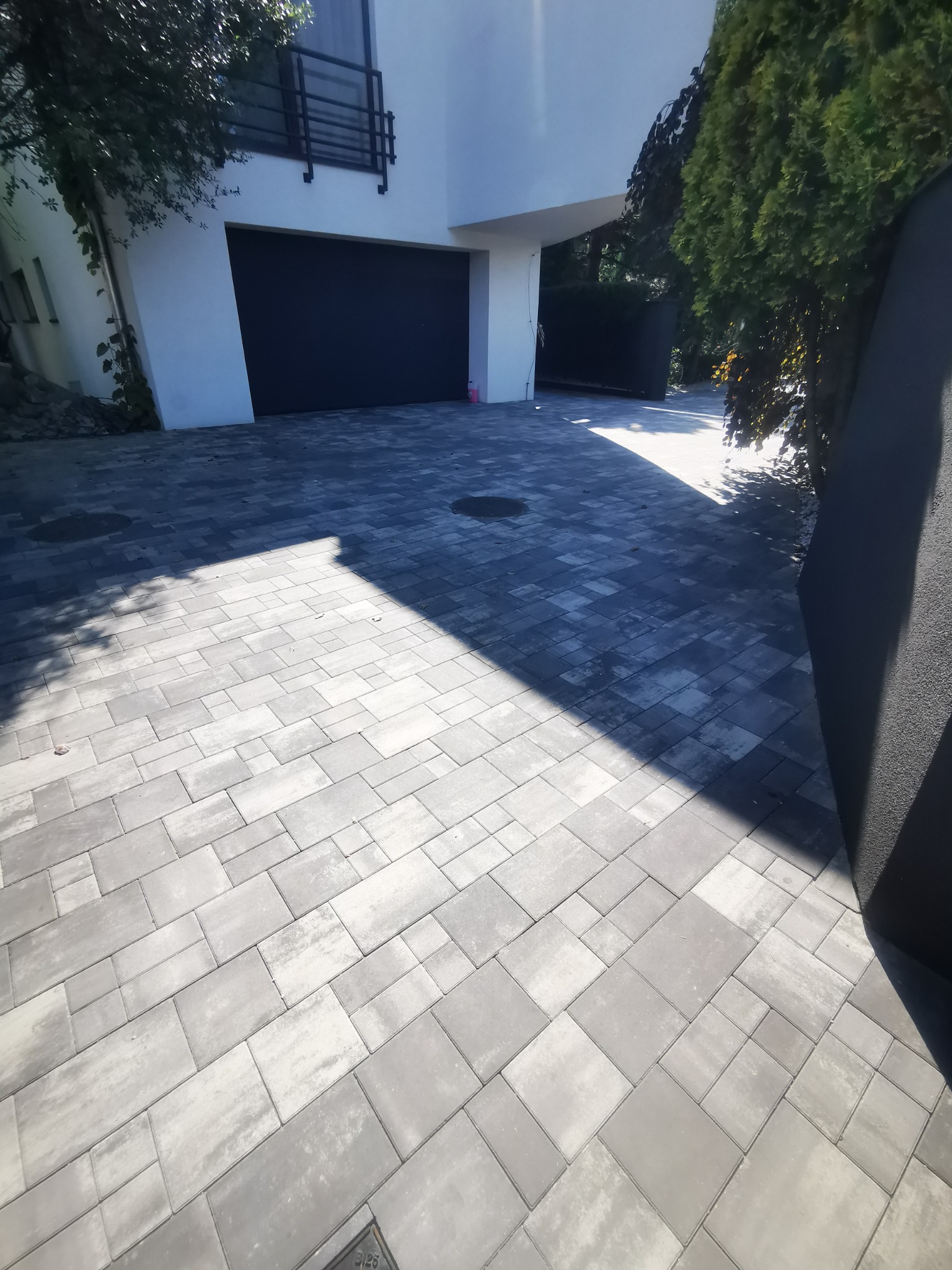 Nowoczesny podjazd z szarej kostki o różnych kształtach, prowadzący do garażu w białym budynku z czarną bramą, częściowo zacieniony przez drzewa.