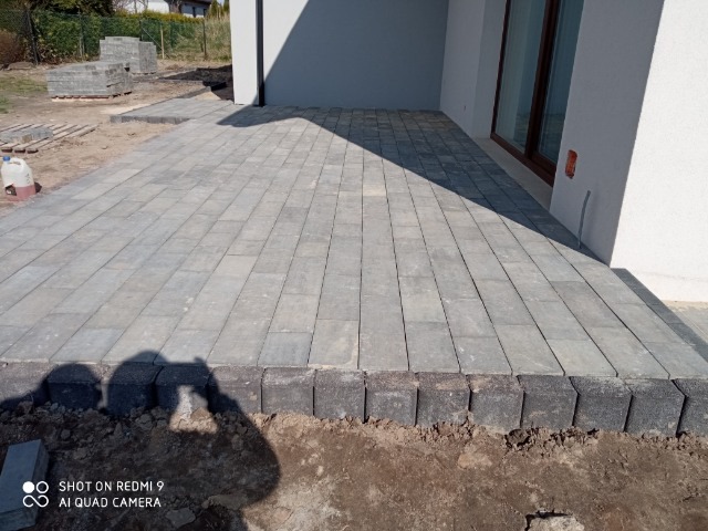 Ułożona szara nawierzchnia z kostki betonowej przed budynkiem, z widocznymi pozostałościami ziemi po pracach brukarskich i stertą kostki w tle.