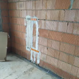 Ściana z pustaków ceramicznych z wyfrezowanymi bruzdami pod instalację elektryczną, widoczne puszki instalacyjne i rurki karbowane prowadzone wzdłuż ściany przy podłodze.