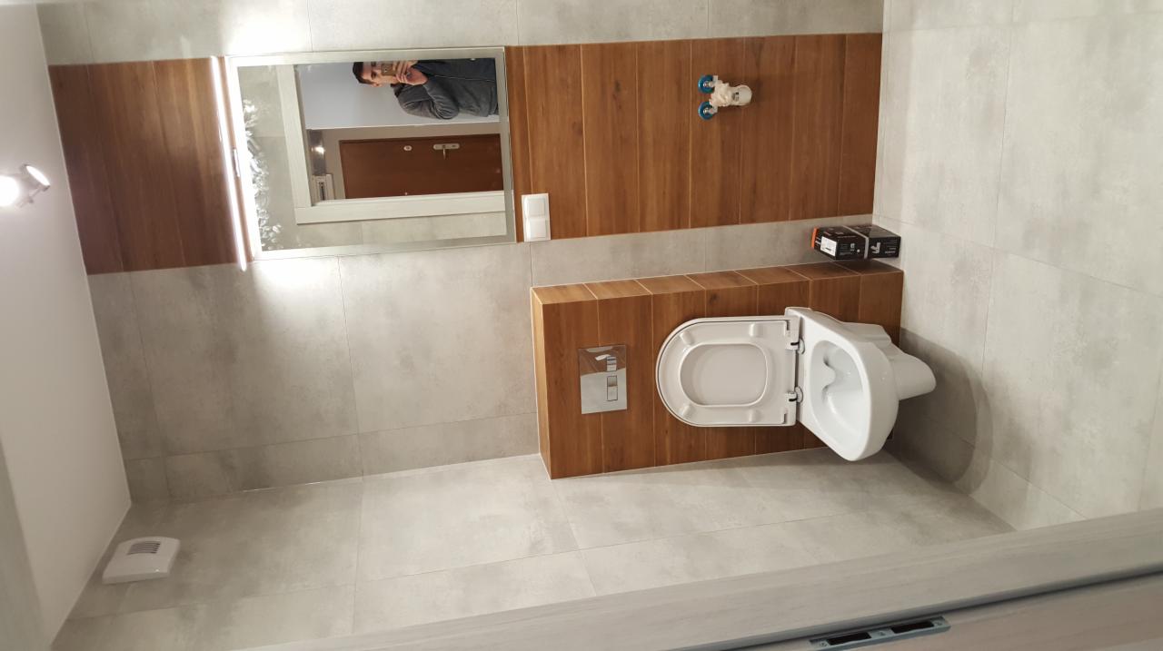 Nowoczesna łazienka z podwieszaną toaletą, drewnopodobnymi panelami na ścianie i lustrem z odbiciem osoby robiącej zdjęcie.