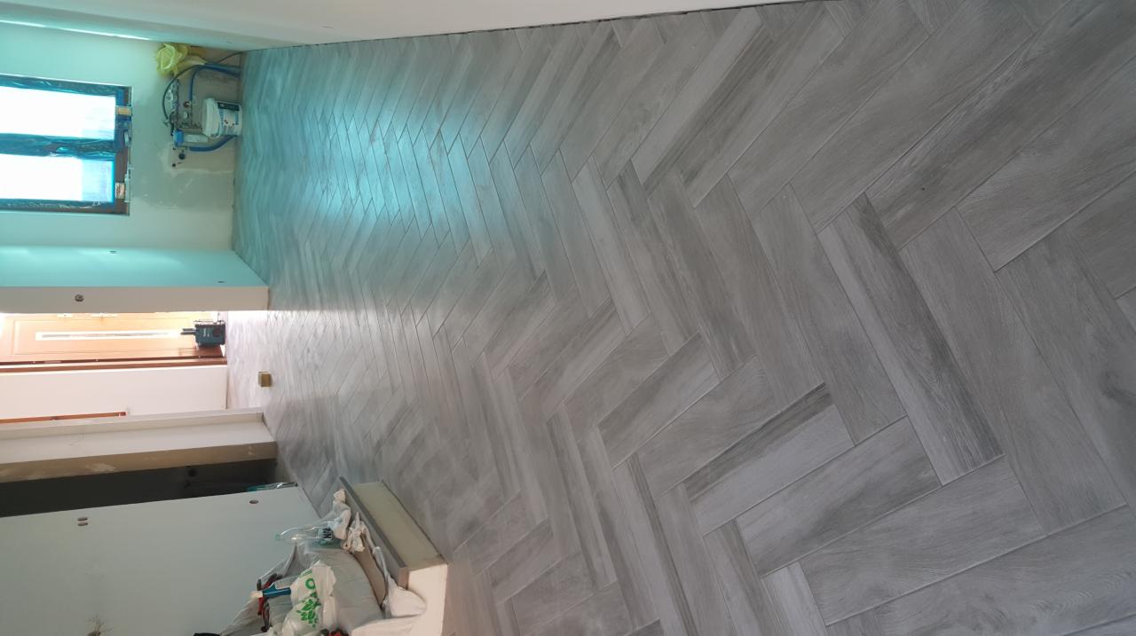 Ułożona w jodełkę szara okładzina ścienna imitująca drewno, z widocznymi instalacjami i oknem w tle, w trakcie wykańczania.