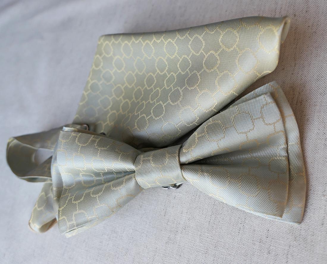 Jasnoszary, jedwabny zestaw: mucha i poszetka z geometrycznym wzorem w odcieniach złota, leżący na kremowym tle.