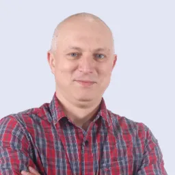 Portret uśmiechniętego mężczyzny w czerwono-niebieskiej koszuli w kratę, z założonymi rękami na tle jasnego tła.