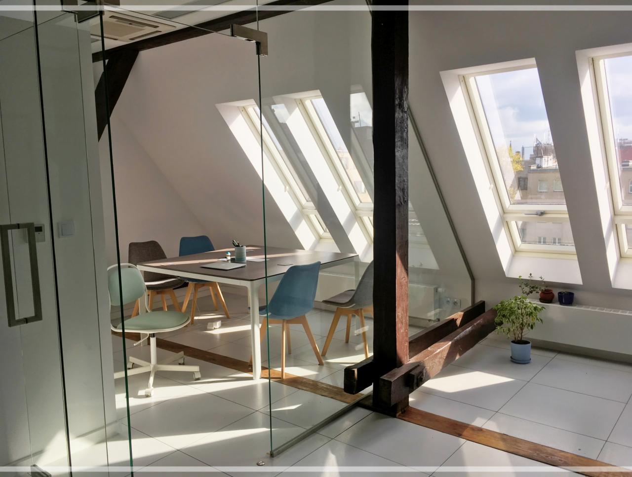 Jasna sala konferencyjna na poddaszu z ukośnymi oknami dachowymi, szklaną ścianą i drewnianą konstrukcją nośną, wyposażona w stół i krzesła.