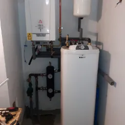 Kompaktowa kotłownia w piwnicy: biały piec gazowy De Dietrich, zbiornik Galmet i czarny hydrofor z manometrami, rury instalacji centralnego ogrzewania i wodnej, narzędzia na stole w rogu.