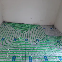 Zielone rury ogrzewania podłogowego ułożone na siatce w surowym stanie, widoczne białe uchwyty mocujące, ściany w trakcie wykończenia.