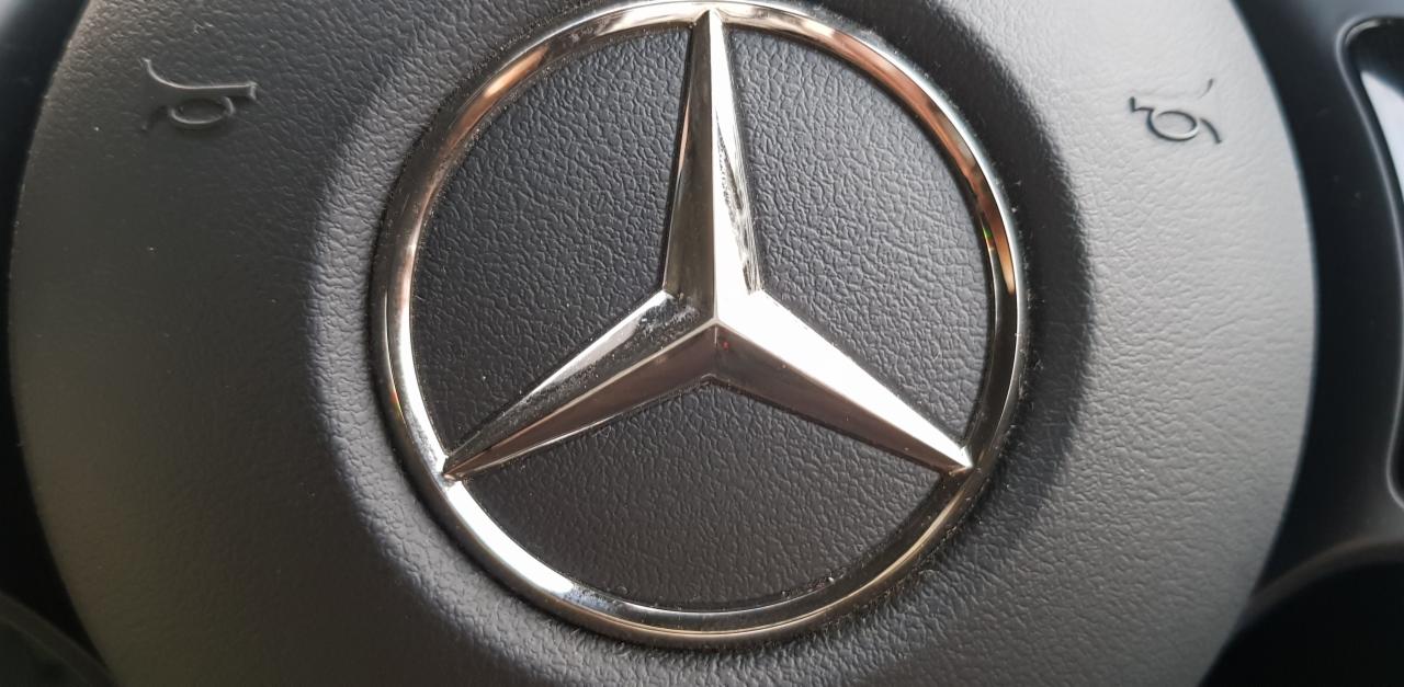 Błyszczące logo Mercedes-Benz na czarnej, teksturowanej kierownicy. Detal z bliska, ukazujący elegancki design i dbałość o szczegóły.