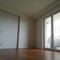 Pokój po malowaniu ścian na jasny odcień, z widocznymi otworami na gniazdka elektryczne, parkietem na podłodze i oknem balkonowym z widokiem na balkon, na którym stoi drabina.