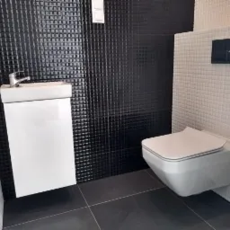 Nowoczesna łazienka z wiszącą toaletą i umywalką na tle czarno-białych, kwadratowych płytek w trakcie wykańczania.