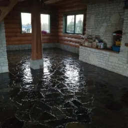 Salon w stylu rustykalnym z podłogą z ciemnego kamienia łupanego, widoczne ściany z kamienia i drewna, kominek oraz schody z bali.