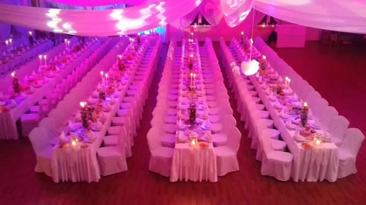 Uroczysta sala bankietowa w różowym oświetleniu: długie stoły z białymi obrusami, krzesła w pokrowcach, świece i dekoracje kwiatowe. Przygotowania do eventu.