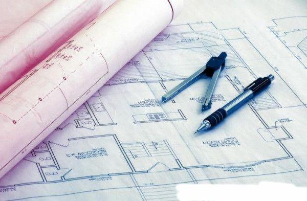 Plan architektoniczny z rzutem mieszkania, rozłożony na stole, z narzędziami kreślarskimi i zwiniętymi planami w tle. Jasne oświetlenie, widok z góry.