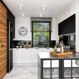 Studio Camilla_Design8 Projektowanie Wnętrz - Nowoczesna kuchnia w bieli i czerni z drewnianą zabudową, marmurową podłogą i wyspą z przeszklonymi szafkami, na blacie butelki wina i kieliszki.