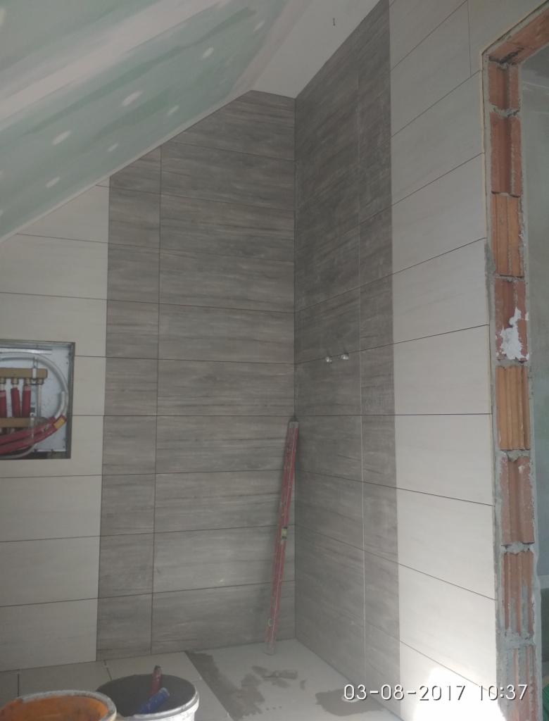 Ściana łazienki w trakcie wykańczania, częściowo wyłożona szarymi płytkami imitującymi drewno, widoczny poziomica oparta o ścianę i odkryta instalacja wodna.