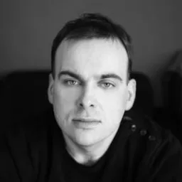 Portret mężczyzny w czerni i bieli, z intensywnym spojrzeniem, na ciemnym tle. Widoczne detale ubrania i rysy twarzy. Delikatne oświetlenie podkreśla strukturę skóry.