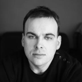 Portret mężczyzny w czerni i bieli, z intensywnym spojrzeniem, na ciemnym tle. Widoczne detale ubrania i rysy twarzy. Delikatne oświetlenie podkreśla strukturę skóry.