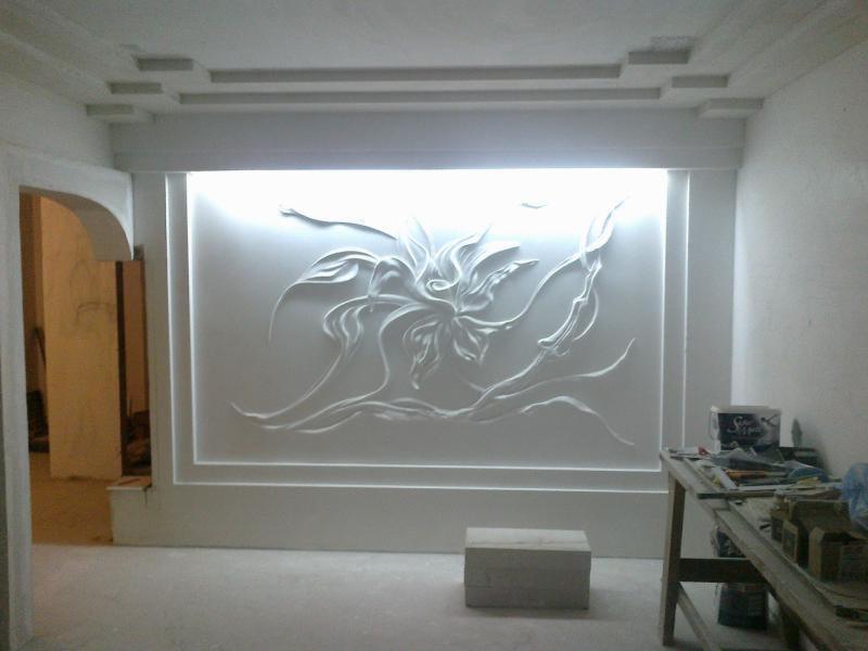 Ściana z gipsową płaskorzeźbą kwiatową podświetloną od tyłu, w surowym wnętrzu z widocznym stolikiem roboczym i materiałami budowlanymi.