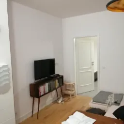 Jasne wnętrze po odświeżeniu, ściany pomalowane na biało, widoczny telewizor na drewnianej komodzie i drewniana podłoga.