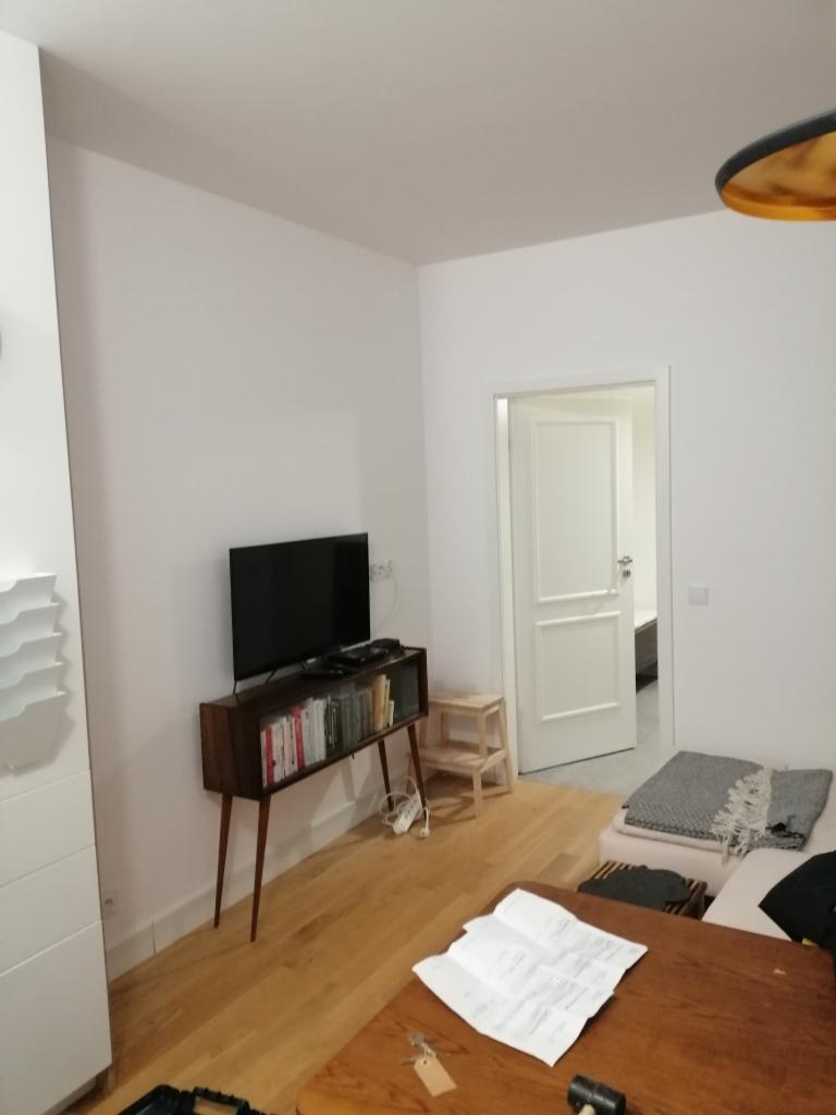 Jasne wnętrze po odświeżeniu, ściany pomalowane na biało, widoczny telewizor na drewnianej komodzie i drewniana podłoga.