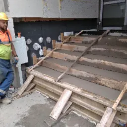 Budowa betonowych schodów wejściowych do budynku; widoczne drewniane szalunki wypełnione świeżym betonem, pracownik w kasku i kombinezonie nadzoruje prace.