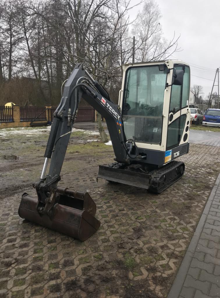 Mała, brudna koparka Terex TC16 na kostce brukowej z błotem, w tle ogrodzenie i drzewa bez liści pod pochmurnym niebem.
