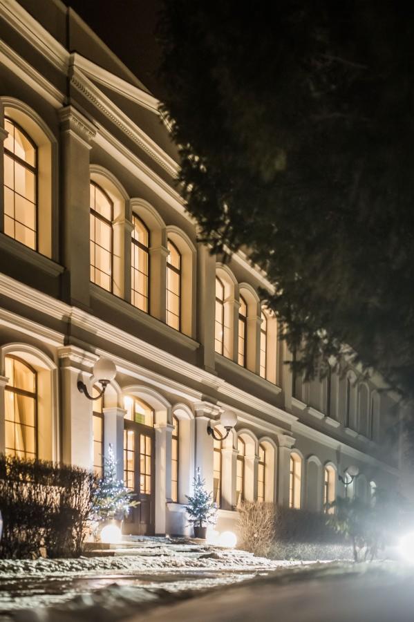 Elewacja budynku z oknami łukowymi oświetlona nocą, dekoracje świąteczne w postaci małych choinek i kulistych lamp przed wejściem, lekko ośnieżony teren.