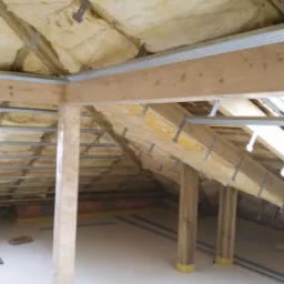 Poddasze w trakcie ocieplania wełną mineralną, widoczne drewniane krokwie z metalowymi mocowaniami, profile metalowe, oraz rolka izolacji ISOVER na podłodze, Śrem.