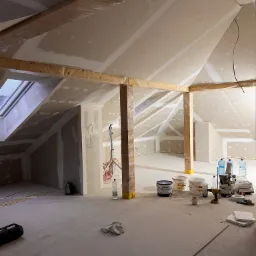 Poddasze w trakcie prac wykończeniowych: widoczne płyty karton-gips na skośnych ścianach, drewniane belki owinięte folią, puszki z farbą, butelki z wodą, wkrętarka i inne narzędzia na podłodze.
