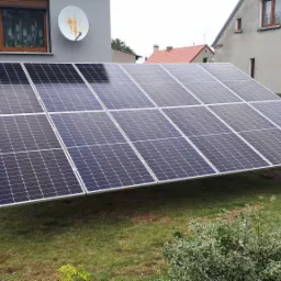 Panel słoneczny ustawiony na trawniku przed domem z szarą elewacją i satelitą, widoczne są także sąsiednie budynki i fragment betonowego ogrodzenia.