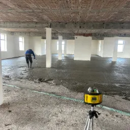 Pracownik w kaloszach rozprowadza szary jastrych na dużej powierzchni w surowym stanie, widoczny laser krzyżowy na statywie, surowy strop z cegieł.