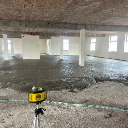 Wnętrze budynku w trakcie prac wylewkowych. Widoczny poziomujący laser na statywie, świeżo wylana masa na podłodze, rury doprowadzające materiał oraz surowy strop z ceglanymi wstawkami.