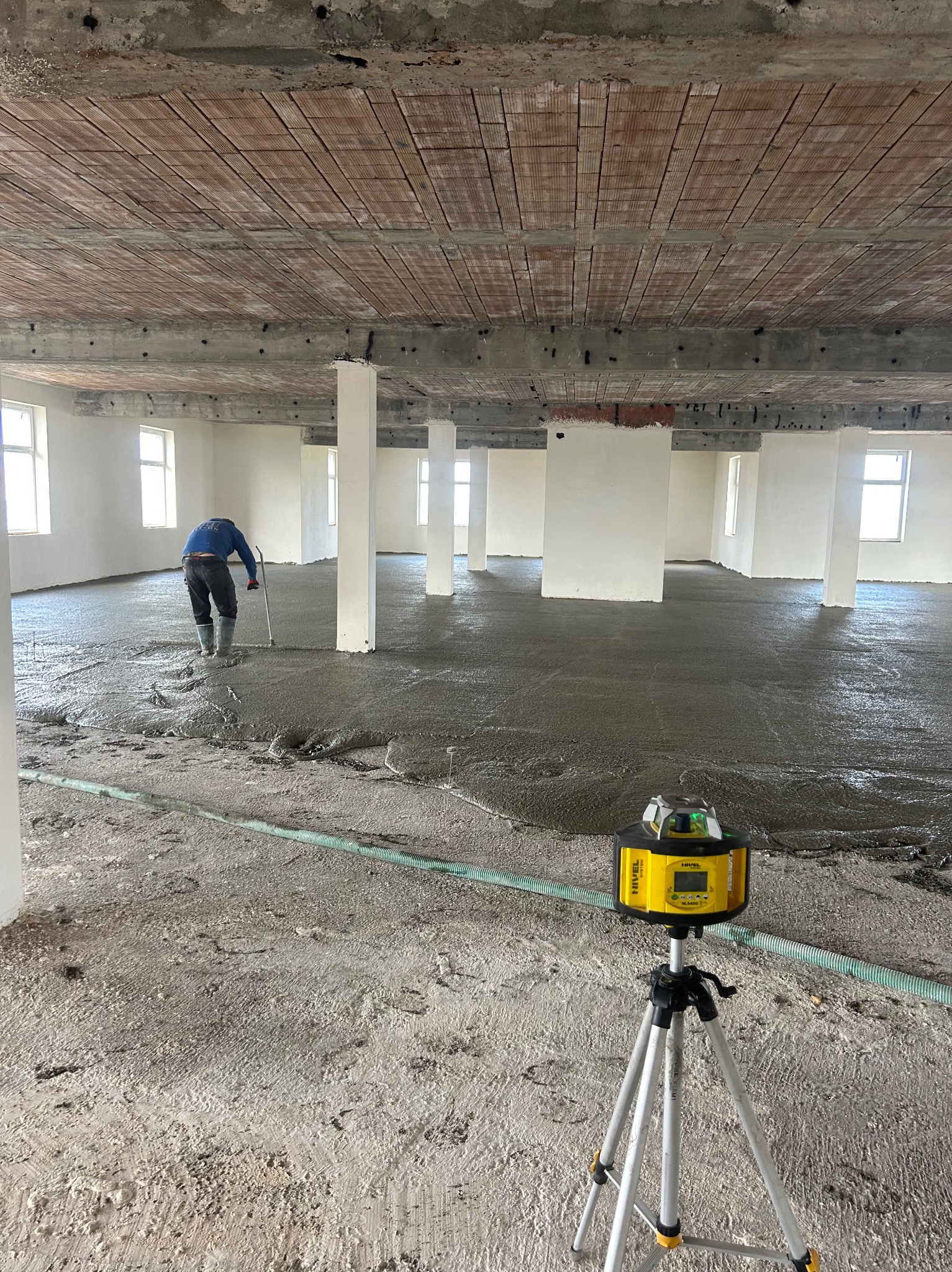 Pracownik w kaloszach rozprowadza szary jastrych na dużej powierzchni w surowym stanie, widoczny laser krzyżowy na statywie, surowy strop z cegieł.