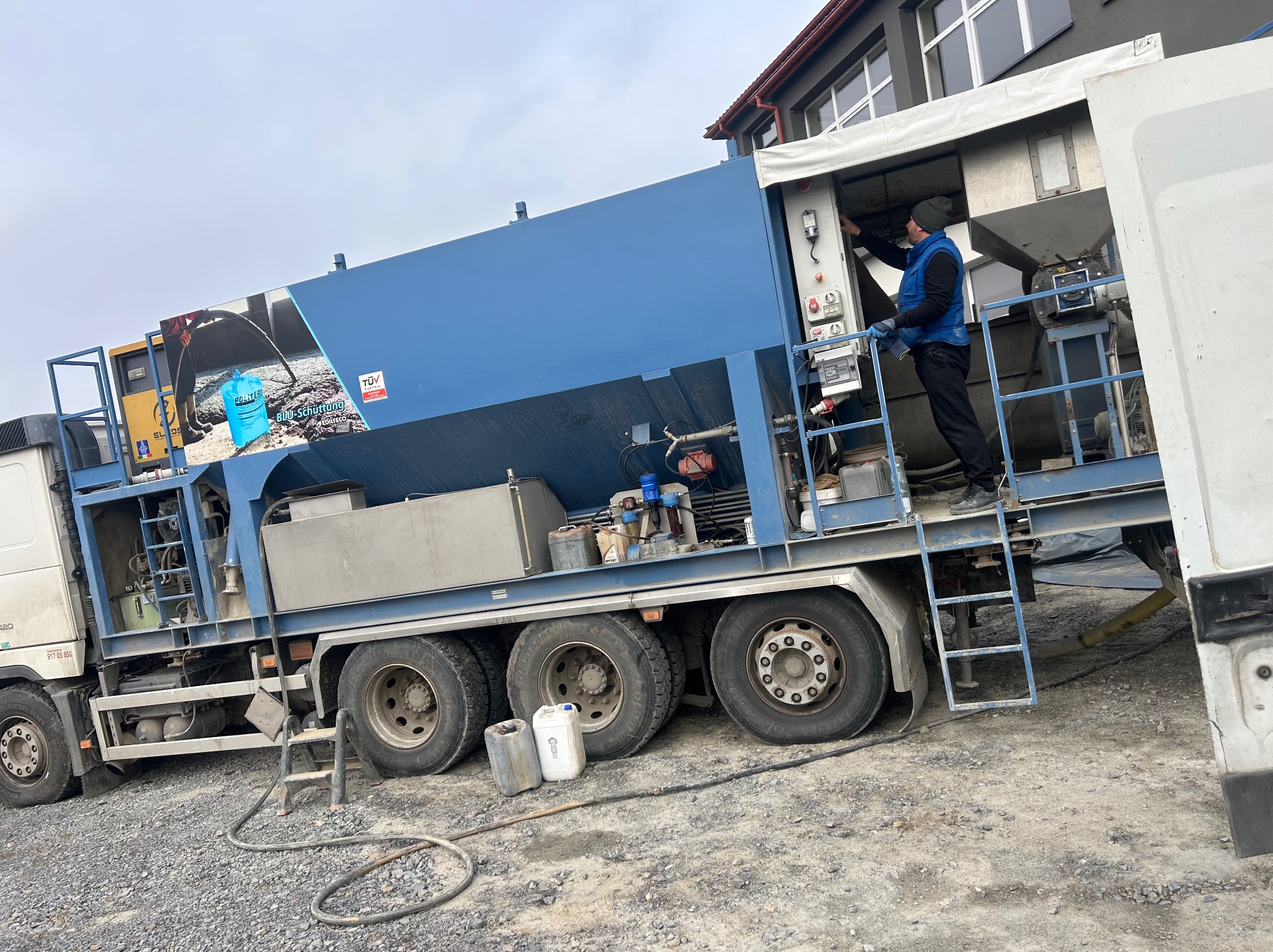 Ciężarówka z niebieską zabudową do przygotowywania wylewek, pracownik obsługuje urządzenie, obok stoją puste pojemniki, na zabudowie reklama worka z wylewką.