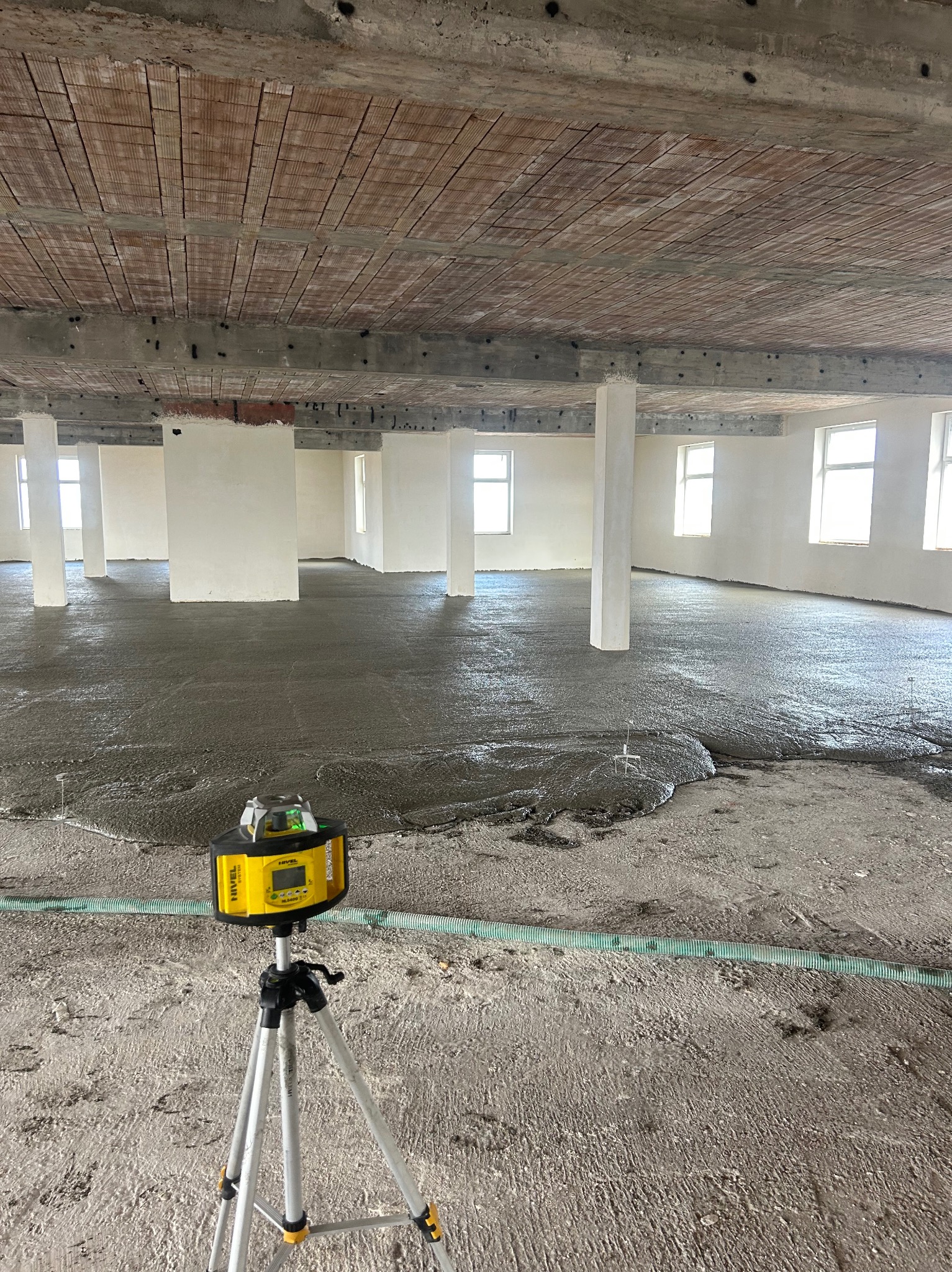 Wnętrze budynku w trakcie prac wylewkowych. Widoczny poziomujący laser na statywie, świeżo wylana masa na podłodze, rury doprowadzające materiał oraz surowy strop z ceglanymi wstawkami.