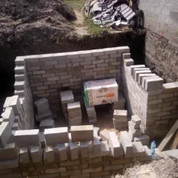 Wykop fundamentów z częściowo wymurowanymi ścianami z bloczków, widoczna sterta ziemi i gruzu w tle, wewnątrz wykopu ułożone bloczki i karton.