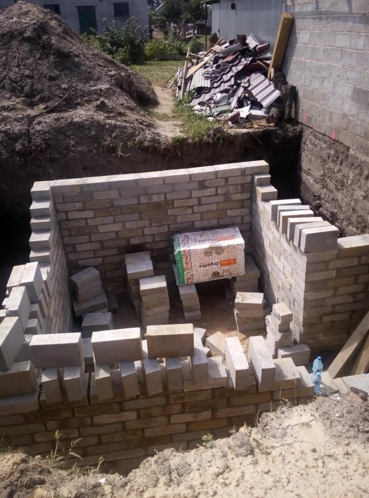 Wykop fundamentów z częściowo wymurowanymi ścianami z bloczków, widoczna sterta ziemi i gruzu w tle, wewnątrz wykopu ułożone bloczki i karton.