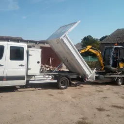 Biały samochód dostawczy z otwartą skrzynią ładunkową, na której przewożona jest mała żółta koparka na przyczepie; widoczny operator w kabinie koparki.