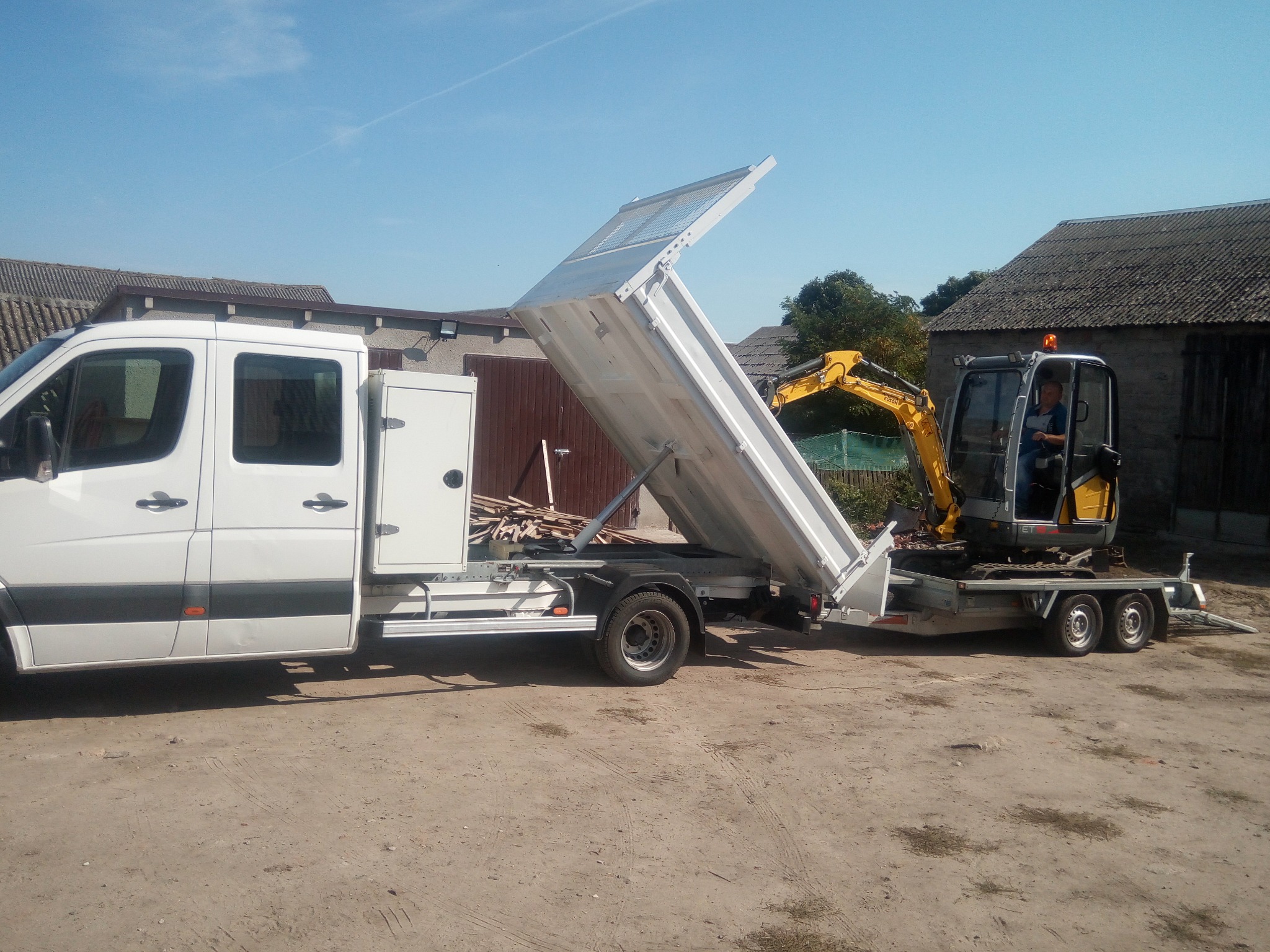 Biały samochód dostawczy z otwartą skrzynią ładunkową, na której przewożona jest mała żółta koparka na przyczepie; widoczny operator w kabinie koparki.