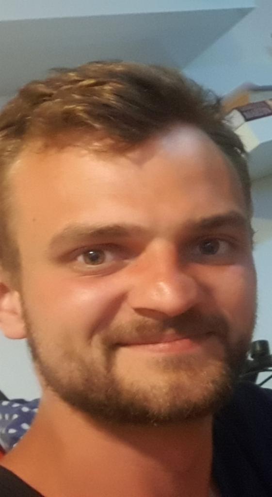 Portret uśmiechniętego mężczyzny z lekkim zarostem, patrzącego wprost w obiektyw, z widocznym fragmentem wnętrza w tle.