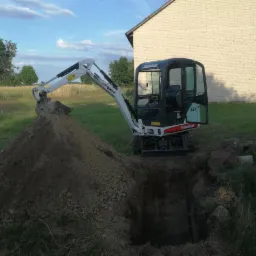 Mini koparka Bobcat 323 podczas prac ziemnych, usypująca kopiec ziemi obok wykopu w trawiastym terenie, z budynkiem w tle.