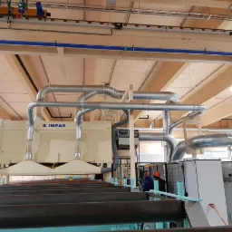 Instalacja przemysłowa z systemem rur wentylacyjnych, widoczne drewniane elementy konstrukcyjne budynku i pracownicy w tle.