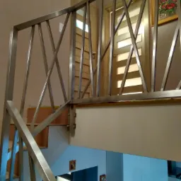 Metalowa balustrada schodowa o geometrycznym wzorze krzyżujących się prętów, widoczna na tle drzwi i ściany z obrazem.