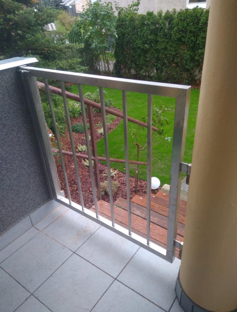 Metalowa furtka balkonowa z pionowymi prętami, widok z bliska na tle zadbanego ogrodu