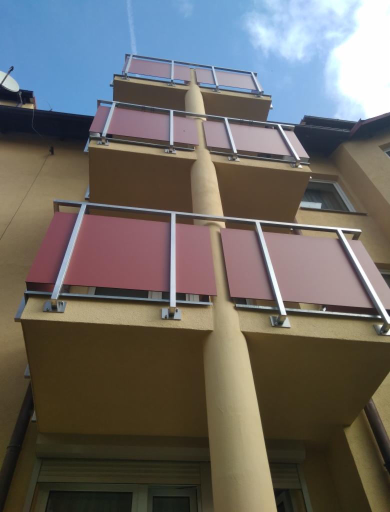 Trzy balkony z metalowymi balustradami i bordowymi panelami, widok z dołu na tle błękitnego nieba.