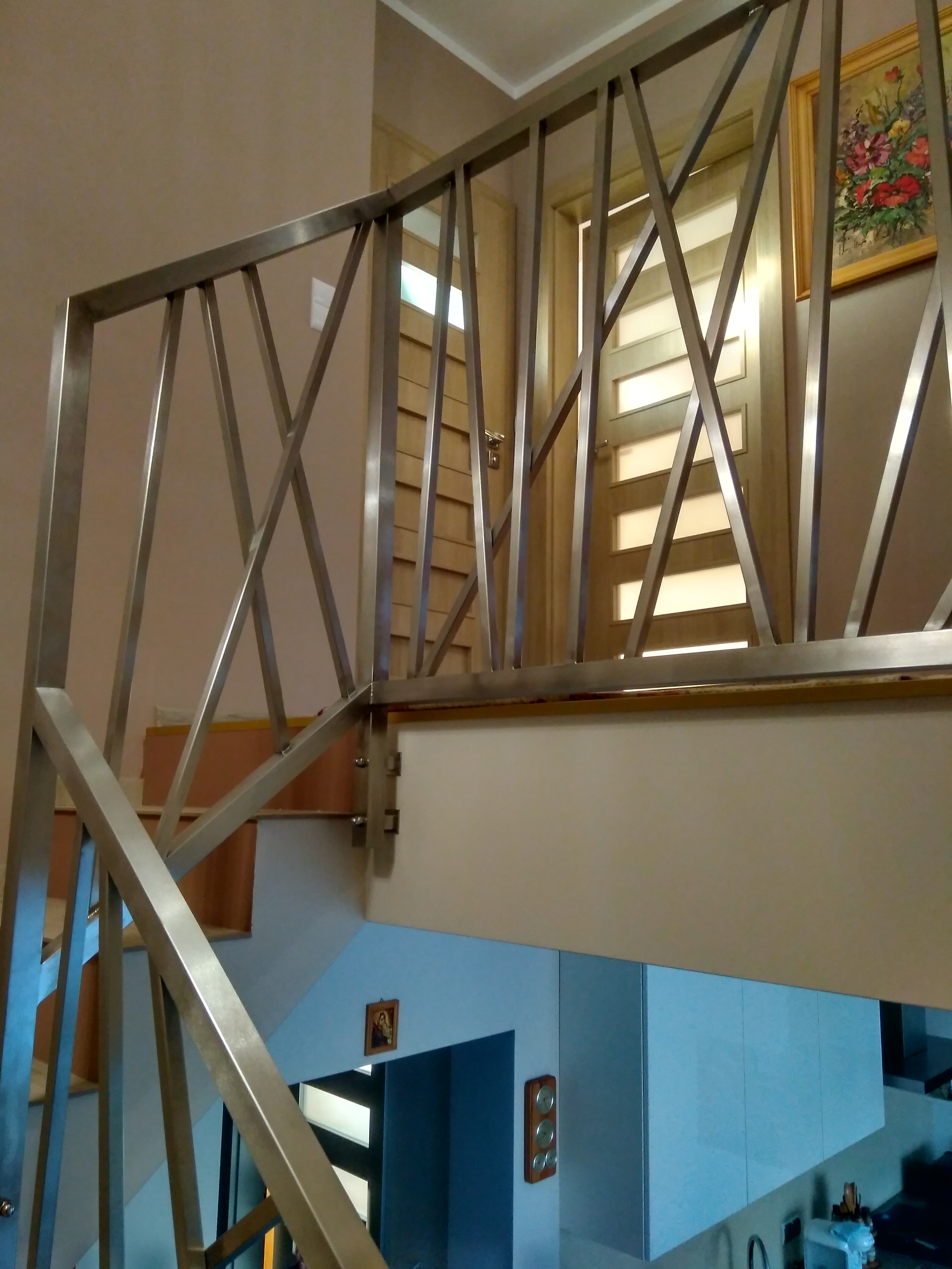 Metalowa balustrada schodowa o geometrycznym wzorze krzyżujących się prętów, widoczna na tle drzwi i ściany z obrazem.