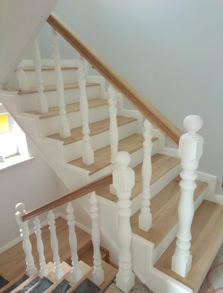 Wnętrze domu: Schody drewniane z białymi balustradami i surową, jasną drewnianą poręczą, widoczne zabezpieczenia na dolnych stopniach.