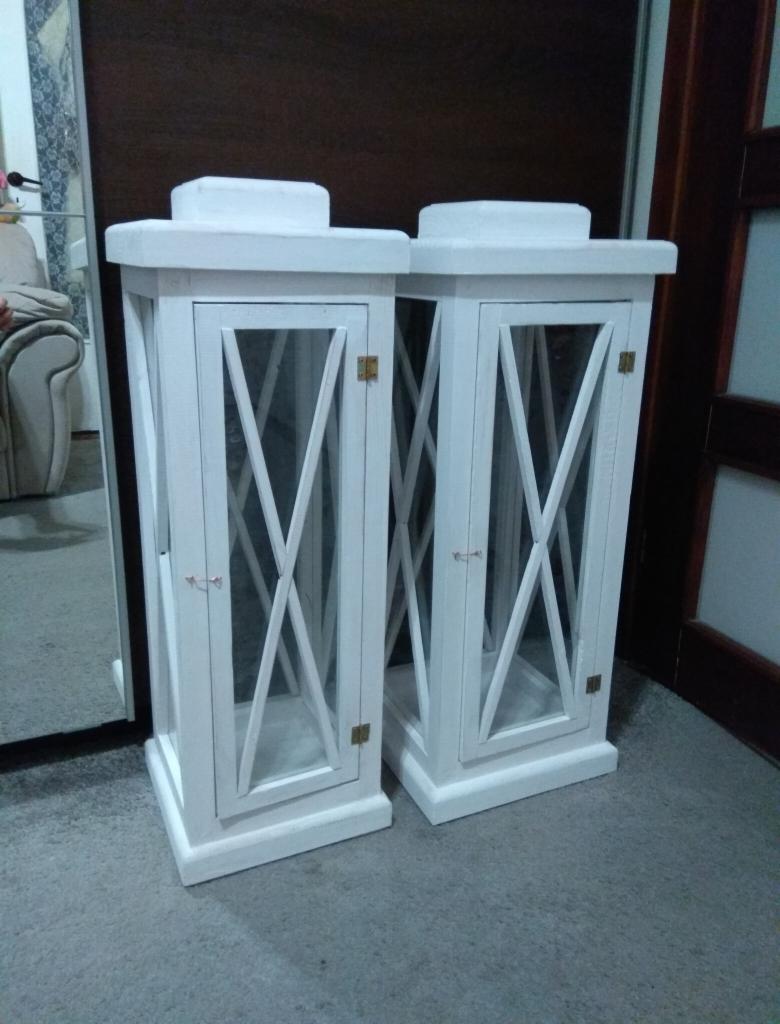 Dwie białe, drewniane latarnie dekoracyjne z przeszklonymi drzwiczkami i krzyżowym wzorem, ustawione na szarym dywanie w pomieszczeniu.