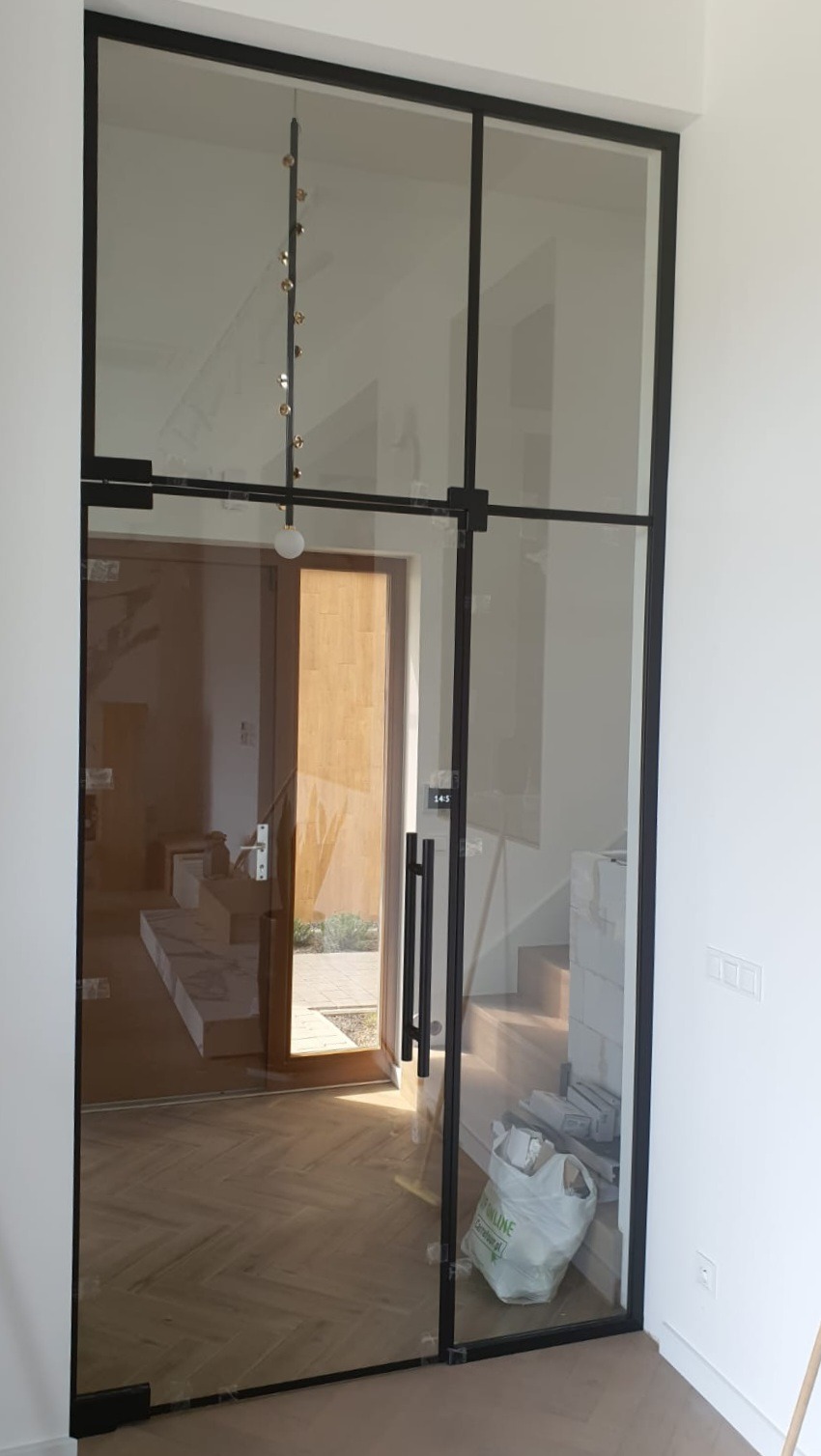 Szklana ściana loftowa z czarnymi profilami oddzielająca wnętrze domu od wejścia, widoczna podłoga z parkietem w jodełkę i schody.