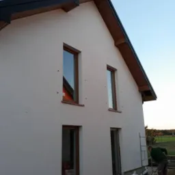 Ocieplona elewacja domu jednorodzinnego z brązowymi oknami, widoczne rusztowanie i pracownik podczas prac wykończeniowych, częściowo ocieplony cokół budynku.